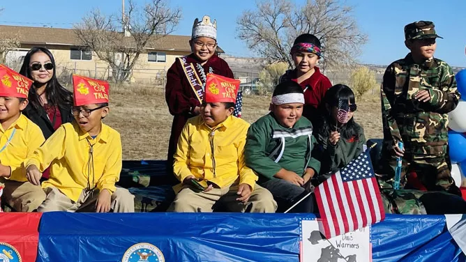Children and adults sit on a decorated parade float during a Veterans Day event. Several children wear yellow shirts and red hats, one holds a small American flag, and another person wears a military-style camouflage outfit. Patriotic decorations, including U.S. military seals and a “Wounded Warriors” sign, are displayed on the float under a clear blue sky.