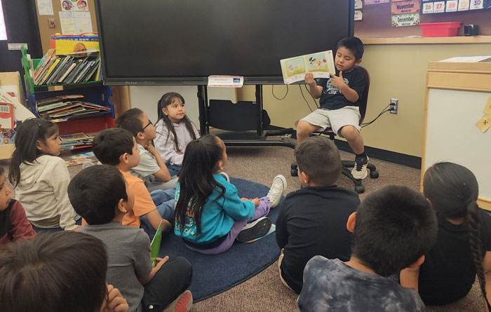A young student sits at the front of a classroom reading a picture book aloud to a group of classmates seated on the floor