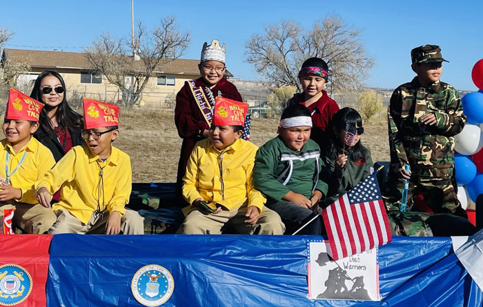 Children and adults sit on a decorated parade float during a Veterans Day event. Several children wear yellow shirts and red hats, one holds a small American flag, and another person wears a military-style camouflage outfit. Patriotic decorations, including U.S. military seals and a “Wounded Warriors” sign, are displayed on the float under a clear blue sky.