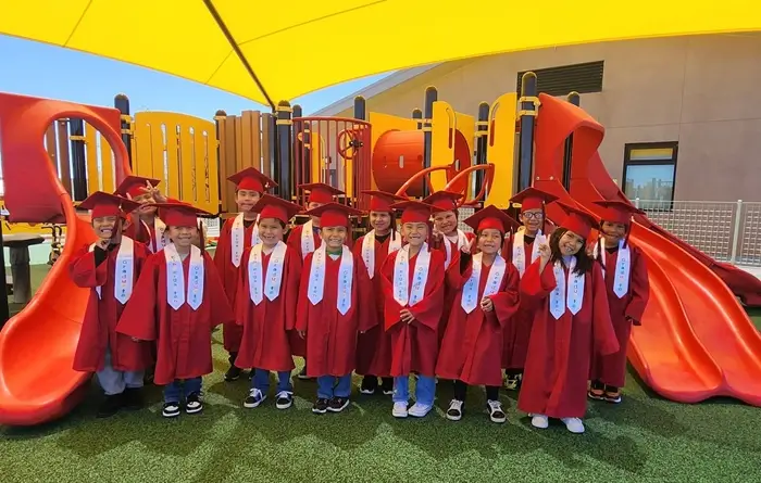 A group of children in red graduation caps and gowns pose together on a playground. 