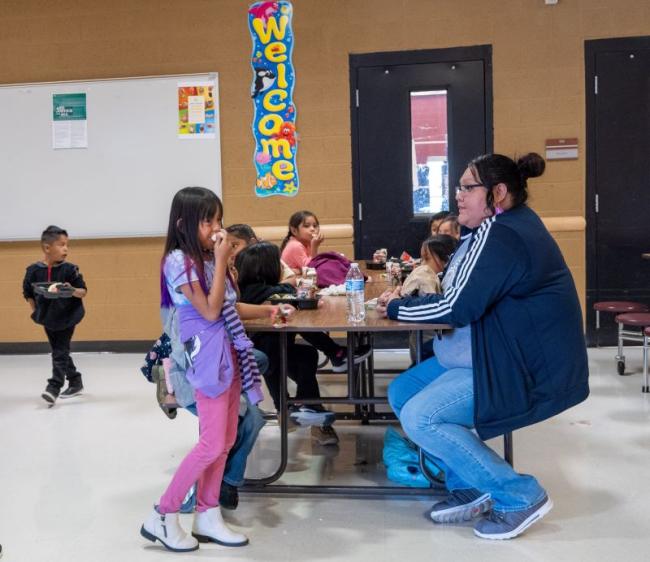 Adult sitting at a full table of students in the cafeteria.