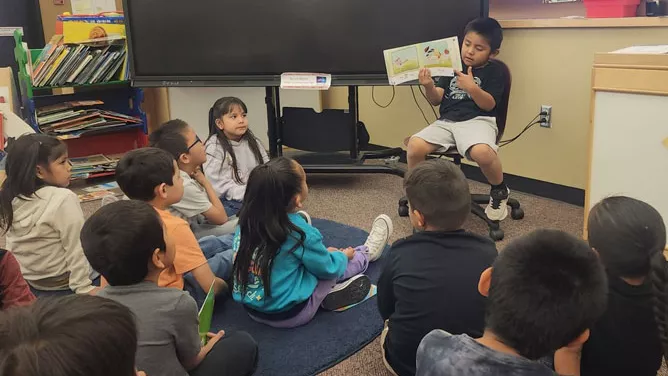 A young student sits at the front of a classroom reading a picture book aloud to a group of classmates seated on the floor