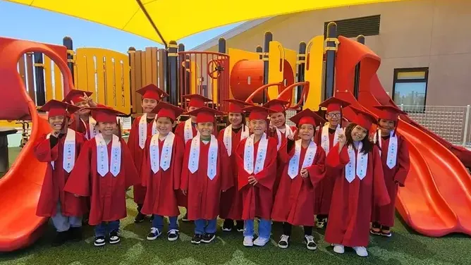 A group of children in red graduation caps and gowns pose together on a playground. 